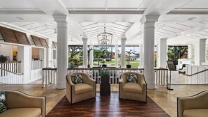 A spacious lobby with high ceilings, white columns, and a central chandelier. There are two beige armchairs facing a glass partition with a view of the outdoors.