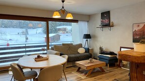 Een gezellige woonkamer met een bank, een salontafel en een eettafel. Er is een groot raam met uitzicht op een besneeerd landschap.