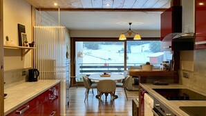 Een moderne keuken met houten kastjes, een eethoek met een ronde tafel en uitzicht op een besneeerd landschap door grote ramen.