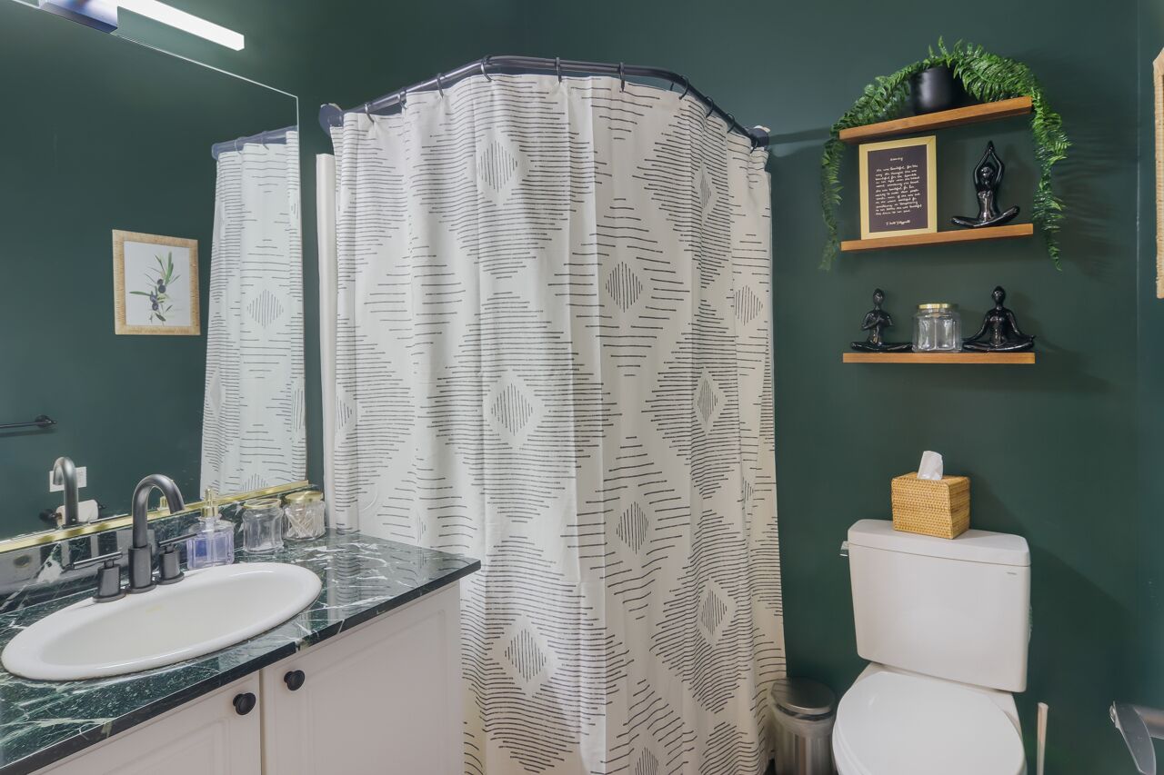 Un baño con un lavamanos blanco, paredes verde oscuro, una cortina de ducha con estampado de hojas y un estante con objetos decorativos.
