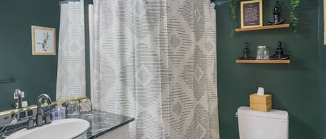 Un baño con un lavamanos blanco, paredes verde oscuro, una cortina de ducha con estampado de hojas y un estante con objetos decorativos.
