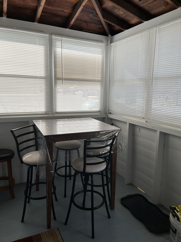 A covered patio with a wooden table and four chairs, large windows with blinds, and a black mat on the floor. - Cute, clean, cozy and pet friendly cottage that is 10 minute walk to the strip. (Geneva)