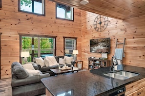 L’intérieur chaleureux de ce chalet est agrémenté de boiseries, d’un coin repas, d’une cuisine équipée d’un évier et de grandes fenêtres offrant une vue sur la verdure.