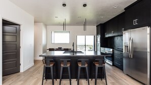 Une cuisine moderne dotée d’un îlot central, de tabourets de bar, de meubles de rangement aux couleurs sombres et d’appareils électroménagers en acier inoxydable.