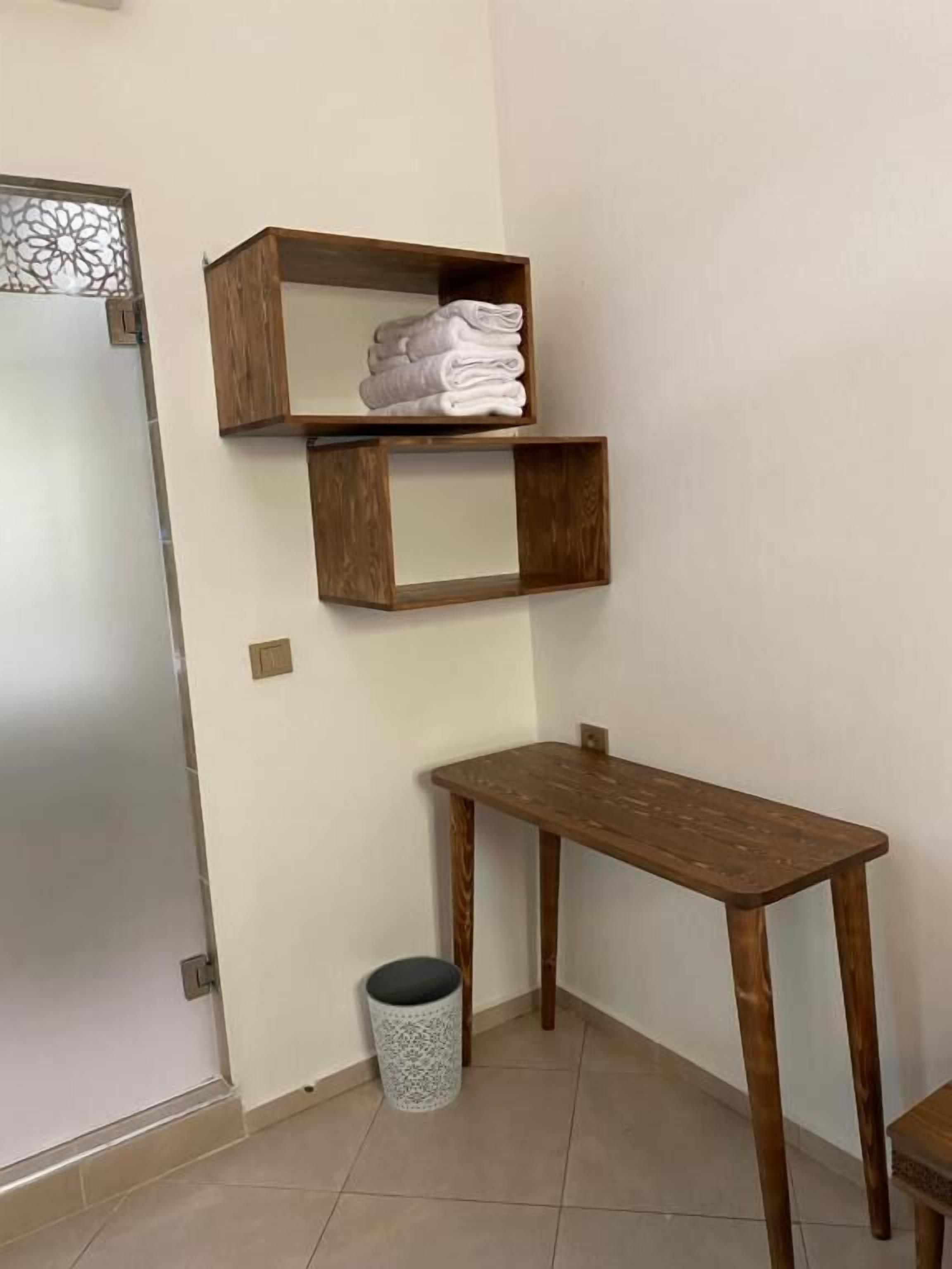 A corner room with a wooden table, a small trash bin, and a shelf with folded towels.