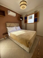 Ein Schlafzimmer mit einem Bett, einem kleinen Holfnachtkästchen, einer Wand-Klimaanlage und einem Fenster mit Blick.