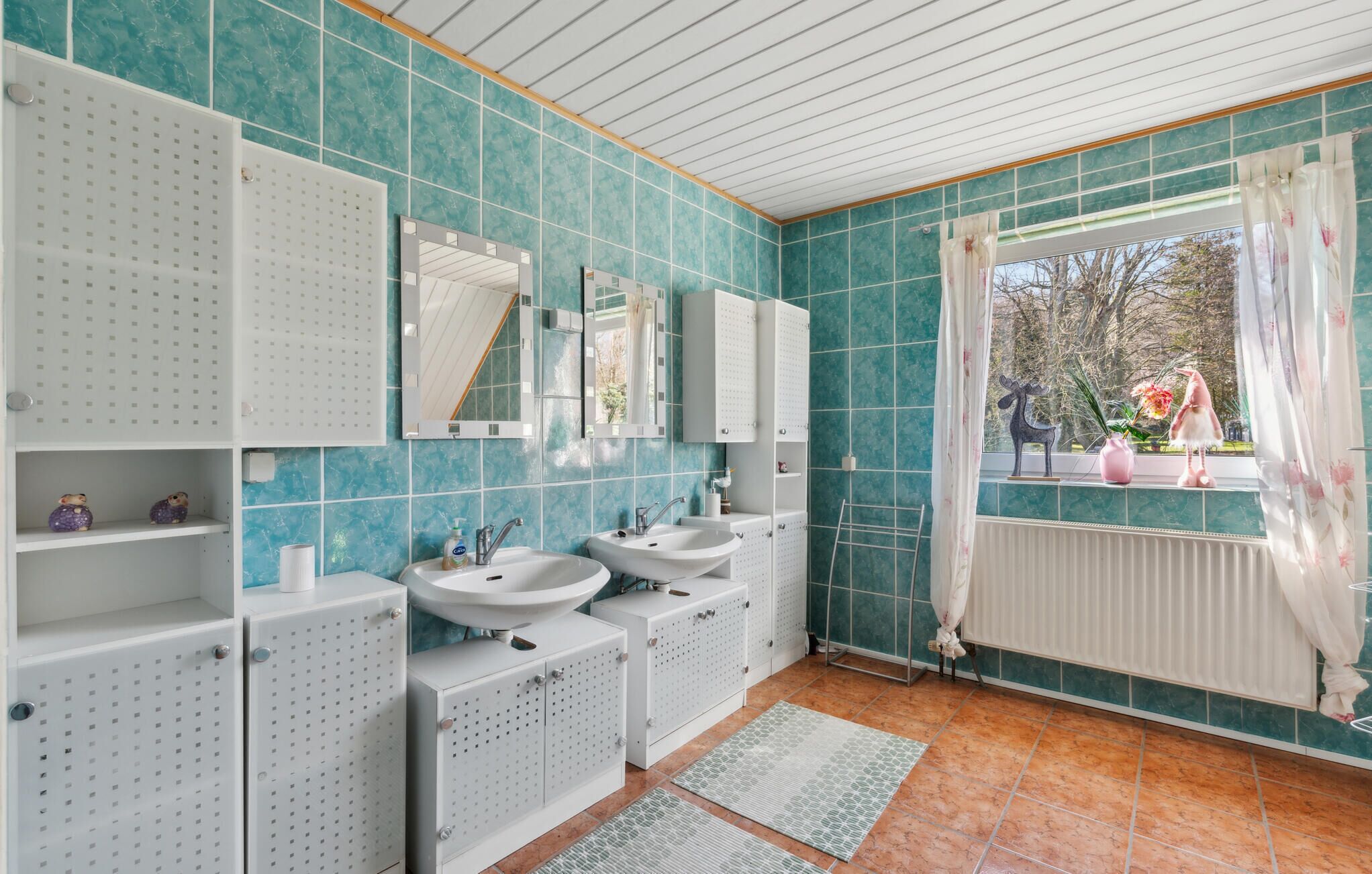 Une salle de bain avec des carreaux de couleur turquoise, deux lavabos, un miroir et une fenêtre donnant sur les arbres.