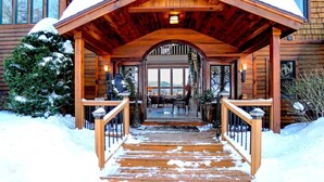 A wooden lodge with a covered entrance, a snow-covered deck, and a view of the interior through a large window.