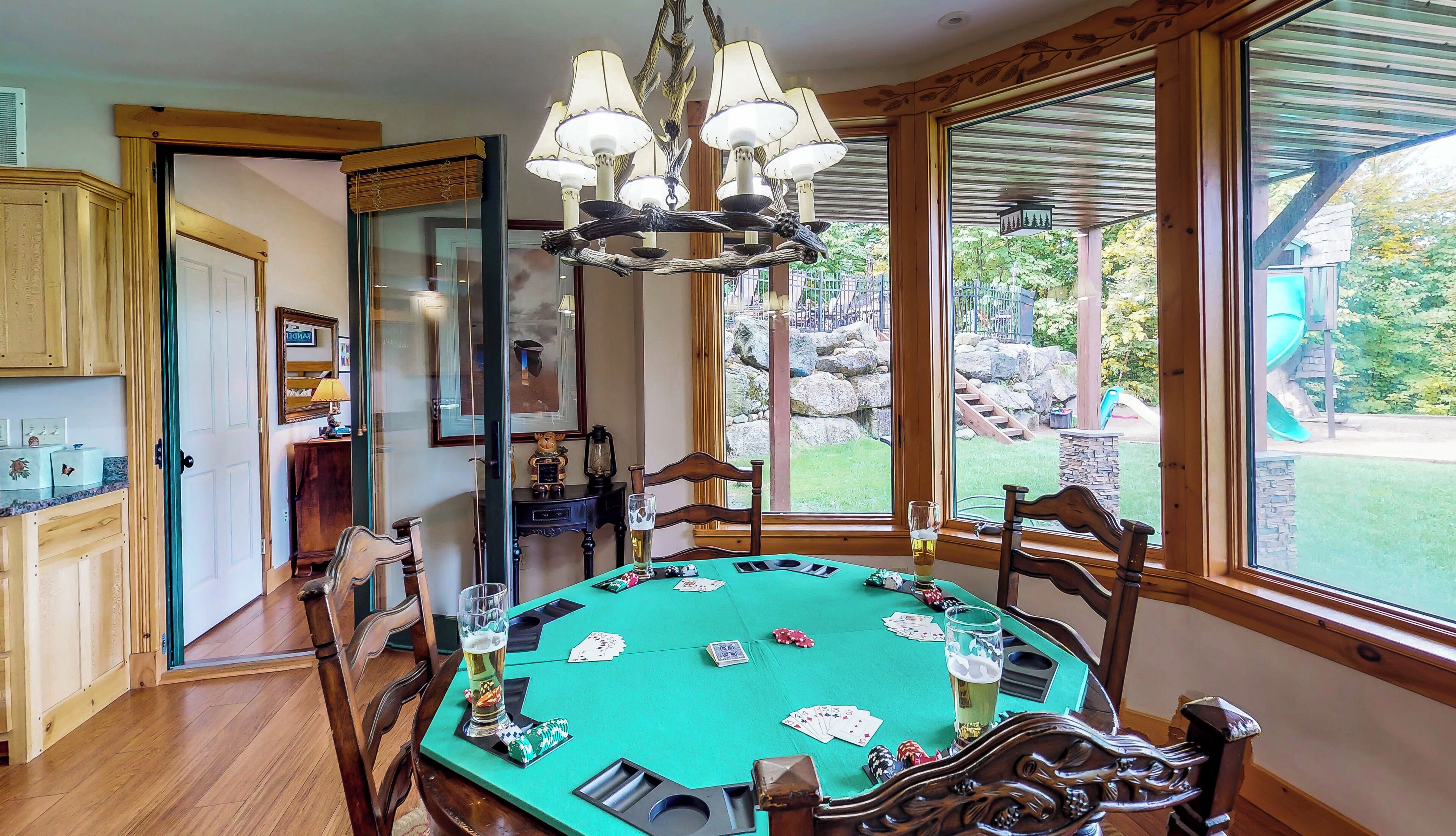Ein Raum mit einem grünen Pokertisch, Holzstühlen, einer Kristallleuchtre und einem großen Fenster mit Blick auf einen Garten und einen Spielbereich im Freien.