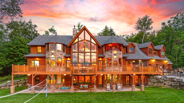 A large, multi-story log cabin with a prominent central glass facade, multiple balconies, and a well-maintained lawn.