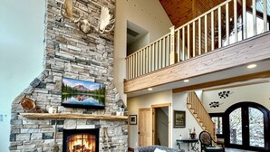 A spacious living room with a stone fireplace, a wooden ceiling, and a balcony.
