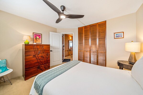 A hotel room with a bed, a desk, a wardrobe, a ceiling fan, and a door leading to another room.
