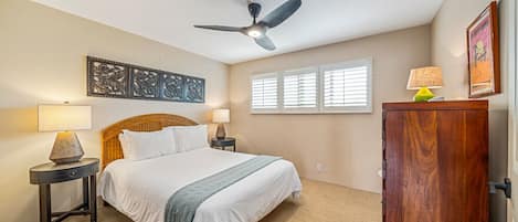 Une chambre à coucher comprenant un lit, deux tables de chevet, un ventilateur de plafond, une fenêtre avec des stores, une armoire en bois et une œuvre d’art accrochée au mur.