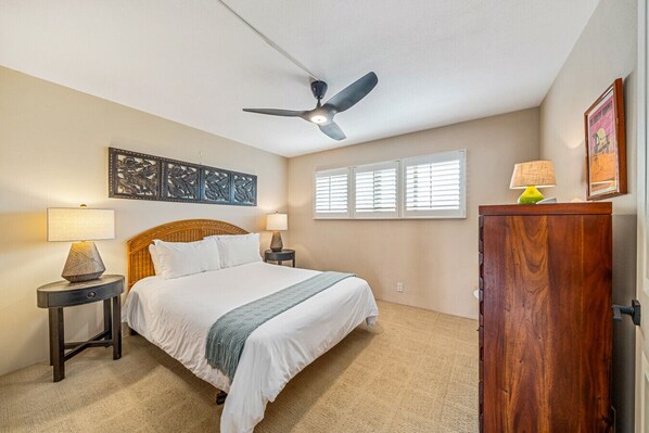 A bedroom with a bed, two bedside tables, a ceiling fan, a window with blinds, a wooden cabinet, and a wall-mounted artwork.