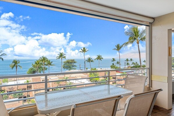 A balcony with a glass table and chairs, overlooking a beach with palm trees and a clear sky.