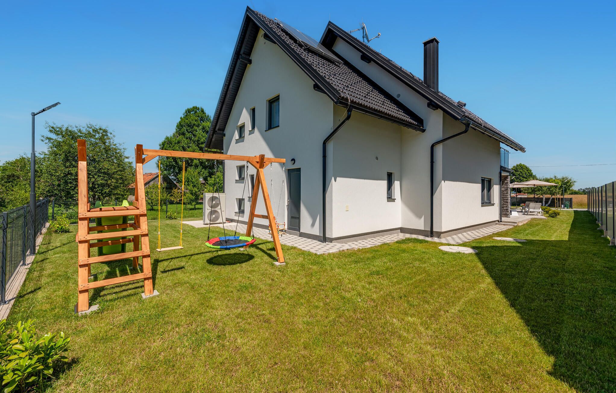 Ein modernes Haus mit weißer Fassade, schwarzem Dach und einem kleinen Garten mit Schaukel und Trampolin.