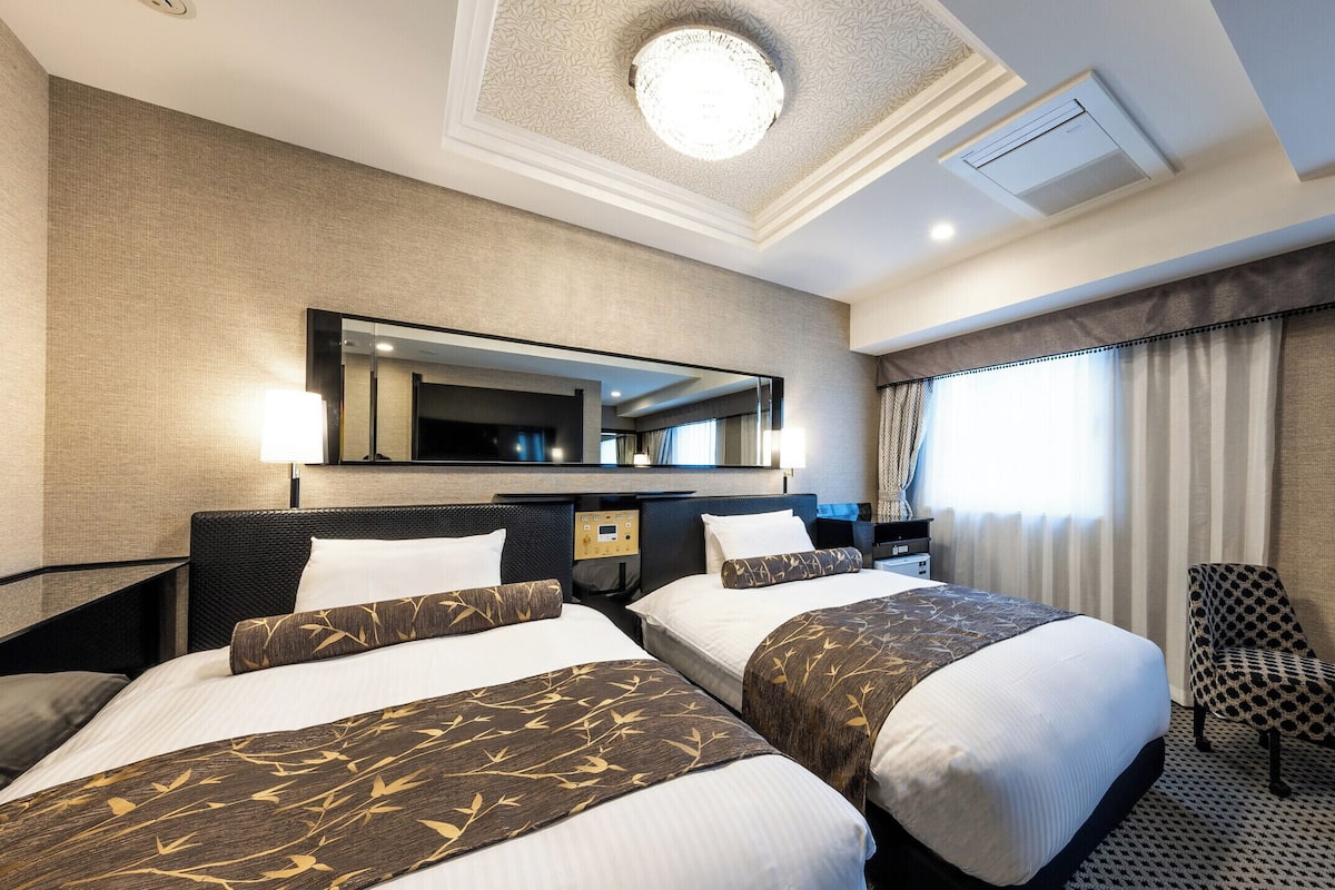 Une chambre d’hôtel avec deux lits, un bureau, une chaise, une télévision et une grande fenêtre avec des rideaux.
