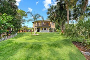 A two-story house with a balcony, surrounded by a well-maintained lawn and tropical vegetation.