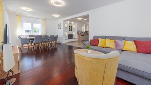 Una sala de estar moderna con un sofá gris, cojines amarillos y rojos, una mesa de centro de madera, y un comedor con sillas grises y una mesa de madera.