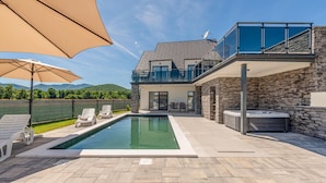Una casa moderna de dos pisos con piscina, mobiliario exterior y un muro de piedra.