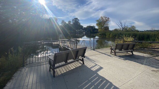 A sunny outdoor area with a paved deck, two wooden benches, and a metal railing overlooking a body of water. - Oasis on Empire near Buckeye Lake! (Thornville)