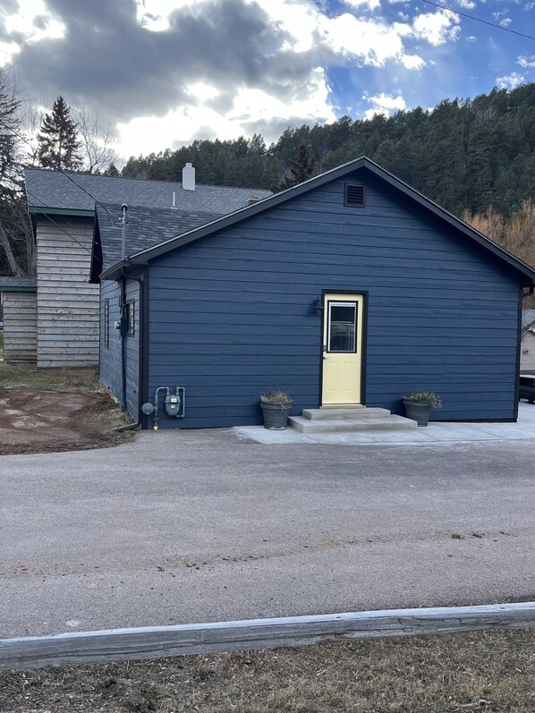 A single-story building with a dark blue exterior, a yellow door, and a small porch area with two potted plants. - “The Yellow Door” perfectly located 2 bedroom 1 bath home, downtown Deadwood (Deadwood)