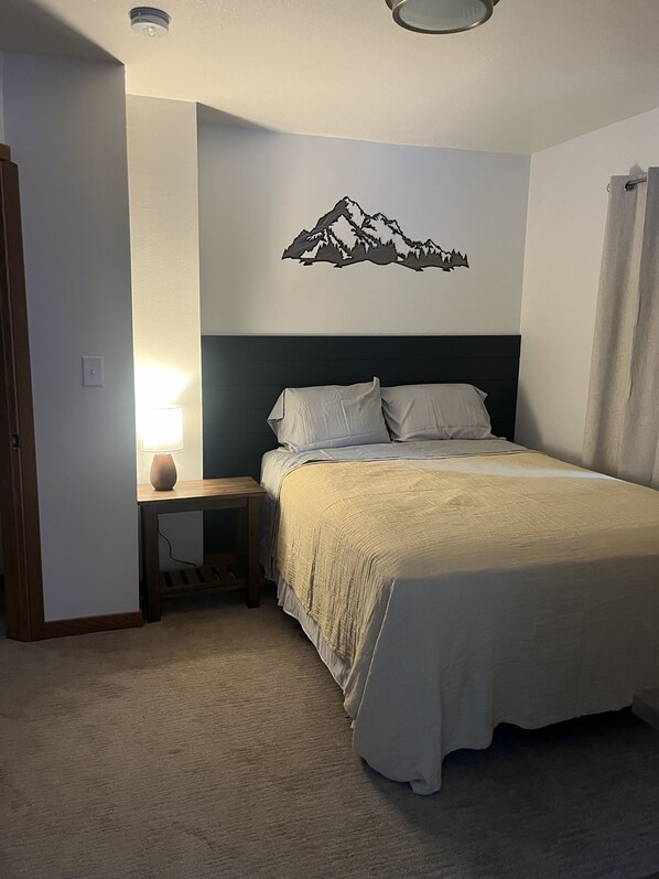 A bedroom with a bed, a nightstand, a lamp, a wall-mounted mountain decoration, and a door.