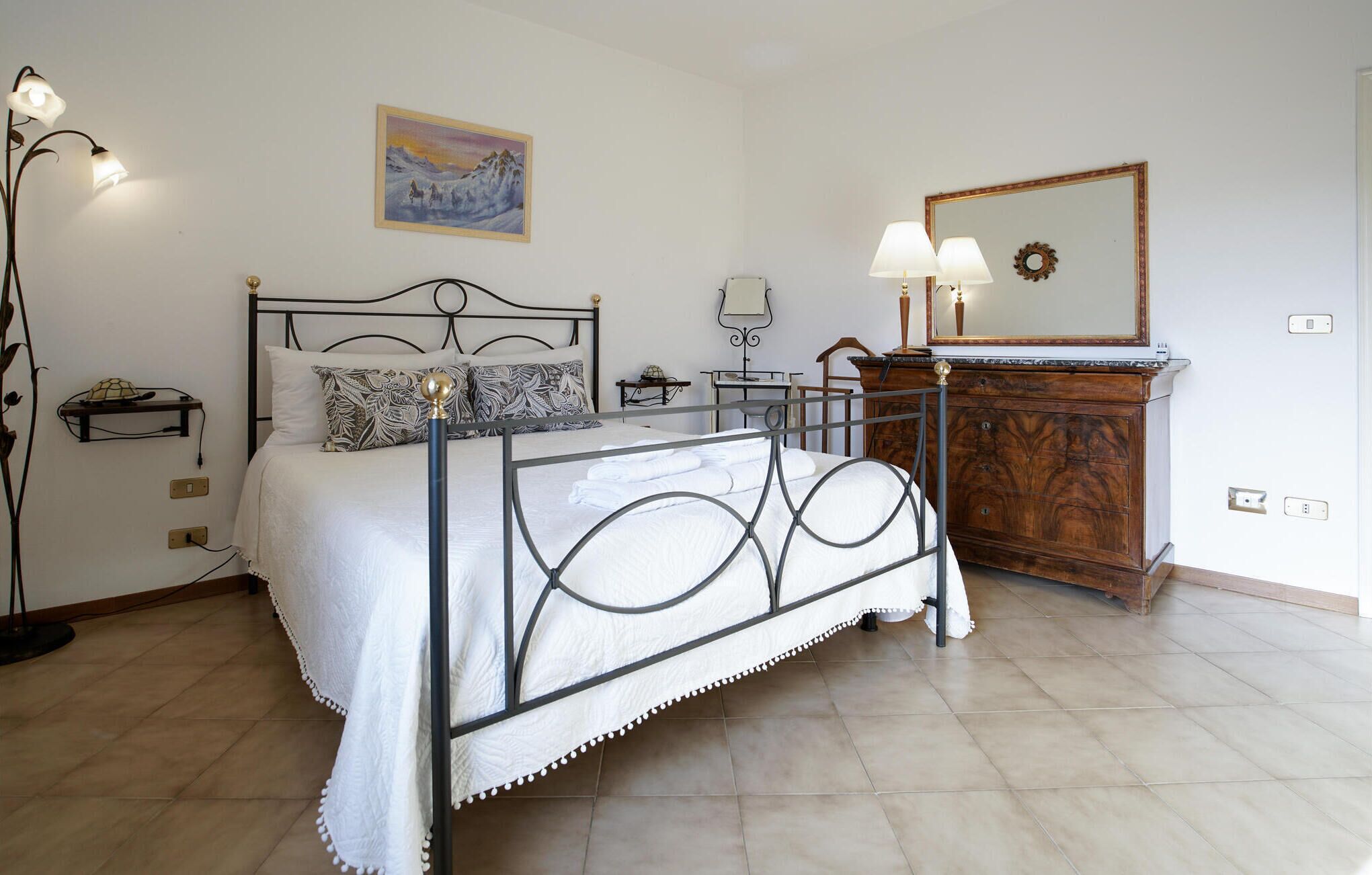 Een slaapkamer met een metalen bedframe, een houten dressoir, een schilderij aan de muur en een spiegel boven het dressoir.