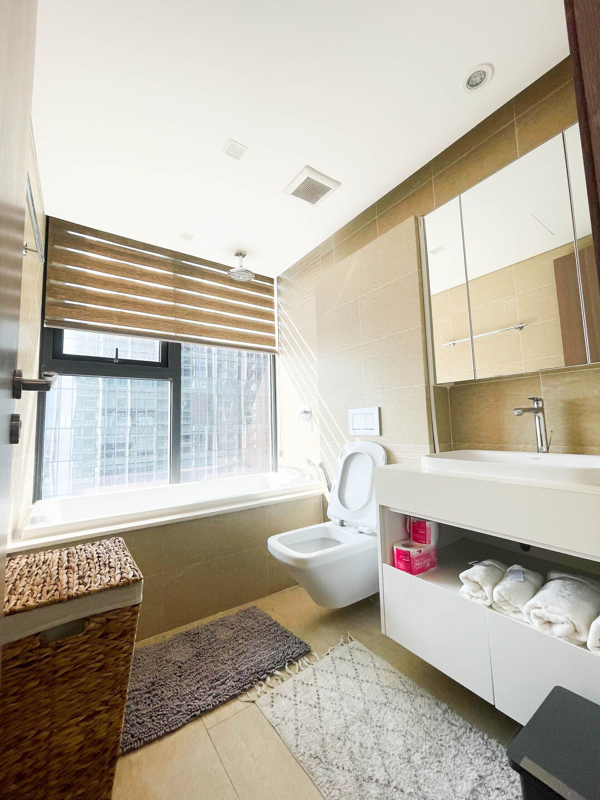 A modern bathroom with a toilet, sink, and large mirror. There is a window with blinds, a rug, and a towel rack.