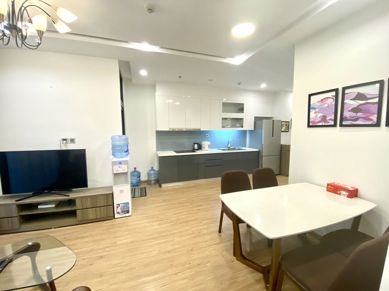 Un salon moderne doté d’un téléviseur à écran plat, d’une table à manger avec des chaises, et d’un coin cuisine avec des placards et un distributeur d’eau.