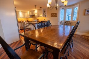A spacious dining area with a large wooden table and six chairs, a kitchen with white cabinets and a window, and a chandelier.