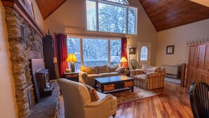Una sala de estar acogedora con chimenea de piedra, pisos de madera y ventanas amplias que ofrecen vistas a árboles cubiertos de nieve.