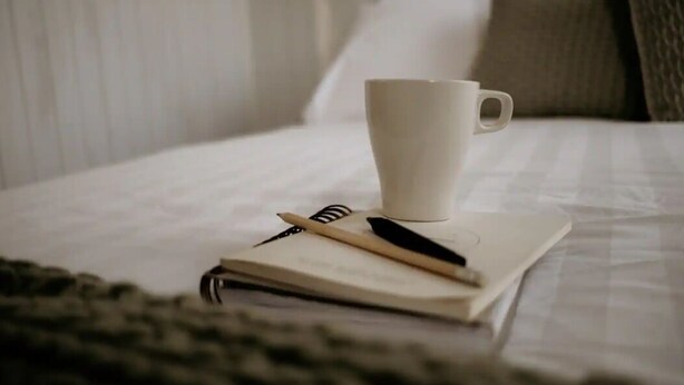 Un lit bien fait, avec un oreiller blanc, une tasse blanche et un carnet sur lequel repose un stylo.