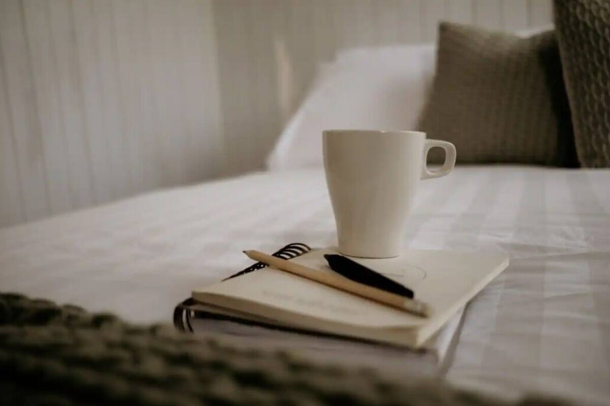 A neatly made bed with a white pillow, a white cup, and a notebook with a pen on it.