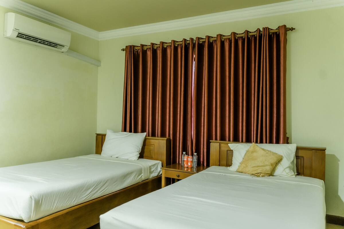 A hotel room with two single beds, wooden headboards, a nightstand with a bottle of Coca-Cola, and a large window with brown curtains.