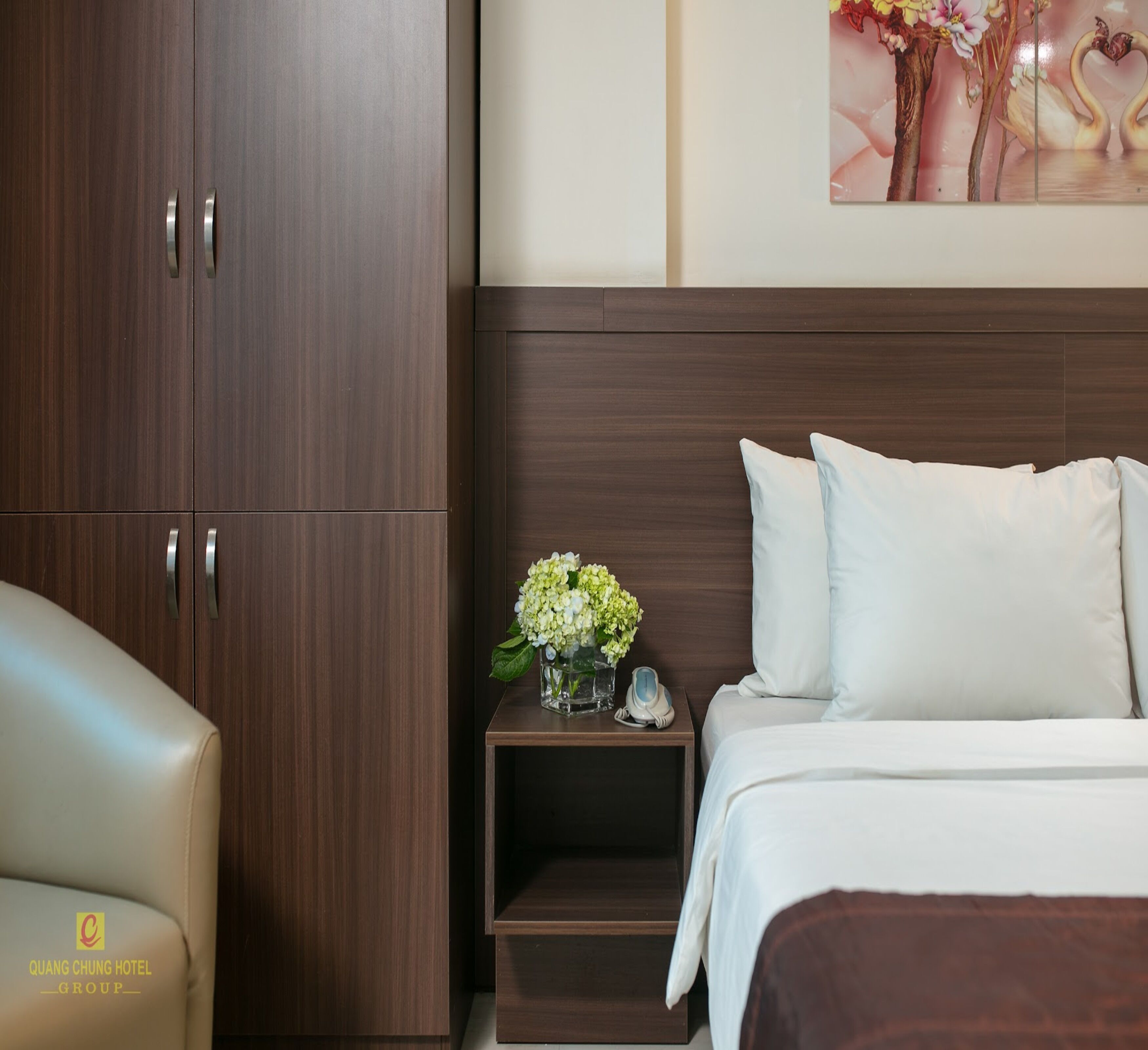 A hotel room with a bed, a nightstand with a vase of flowers, and a wooden wardrobe.