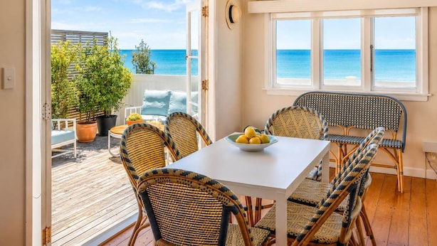 A room with a white table, wicker chairs, a view of the sea, and a balcony with plants. - Beachfront Bliss - Opposite Onetangi Beach (Waiheke Island)