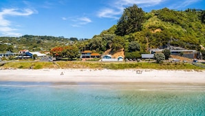 A coastal beach with clear turquoise waters, a sandy shore, and a backdrop of lush green hills with buildings. - Beachfront Bliss - Opposite Onetangi Beach (Waiheke Island)
