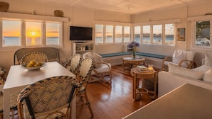 A room with a dining table, chairs, a television, and large windows overlooking the sea. - Beachfront Bliss - Opposite Onetangi Beach (Waiheke Island)