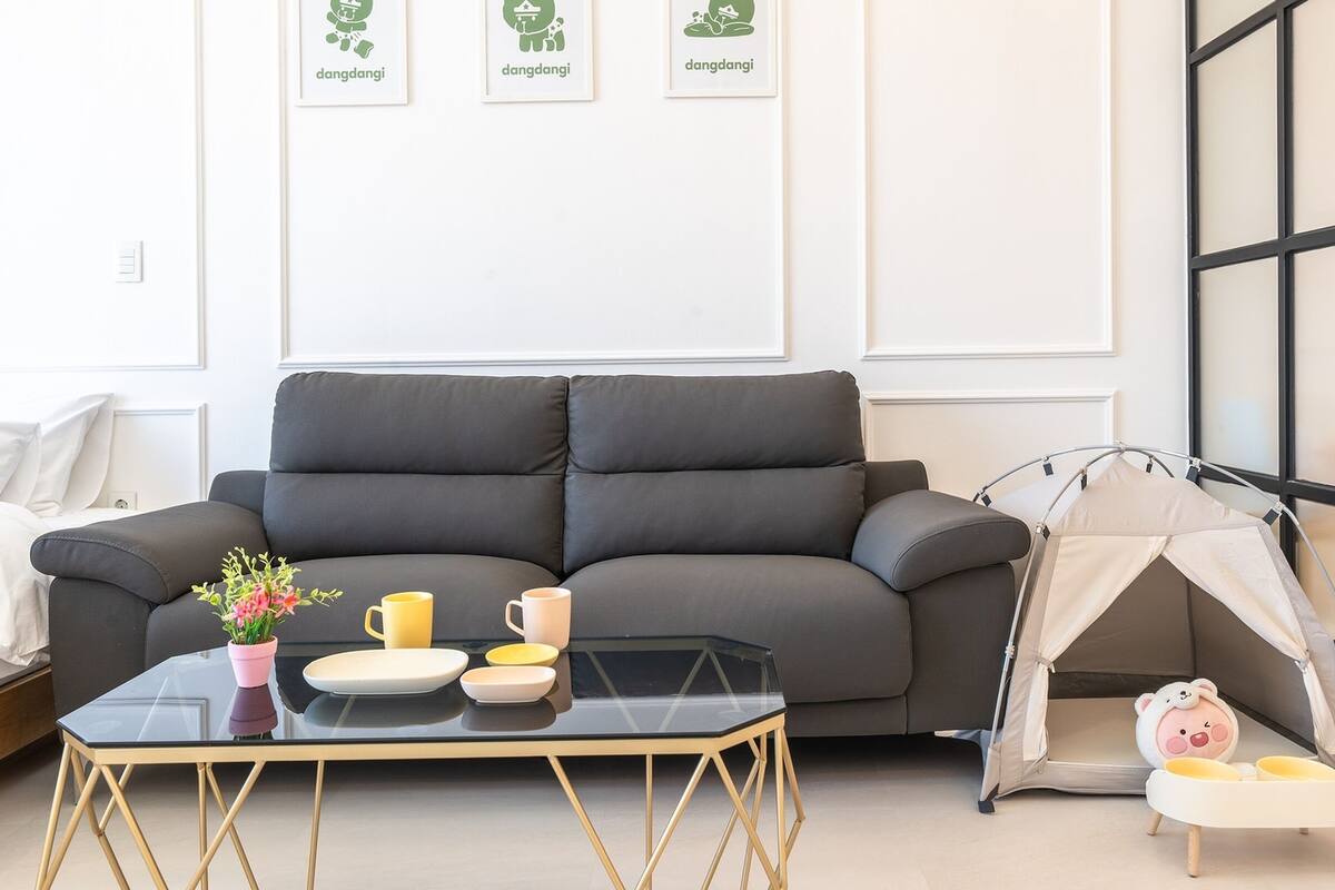 A modern living room with a grey sofa, a glass coffee table with yellow and white plates, a pink vase with flowers, and a tent-like structure with a toy sheep.