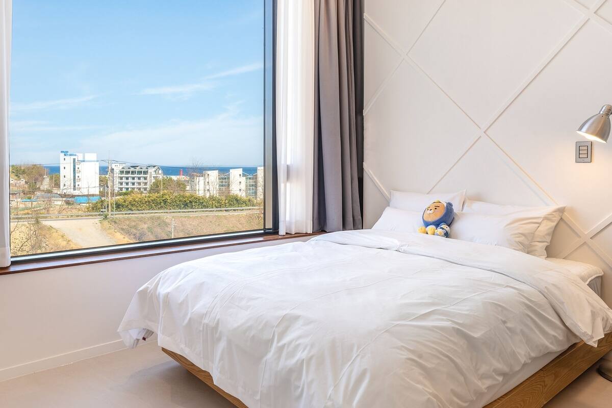 A bedroom with a large window, white bedding, and a plush toy on the bed.