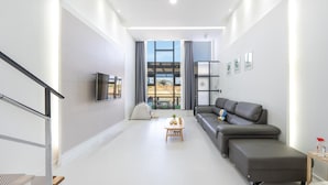 A modern living room with a sofa, a TV mounted on the wall, a small table with decorative items, and a view through a large window. - Gangneung Dangdangi Dog Pension Blue (Gangneung)