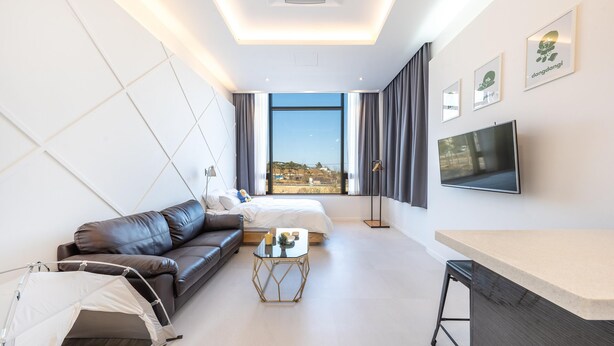 A modern hotel room with a large bed, a sofa, a small table, a TV, and a view of the city. - Gangneung Dangdangi Dog Pension Blue (Gangneung)