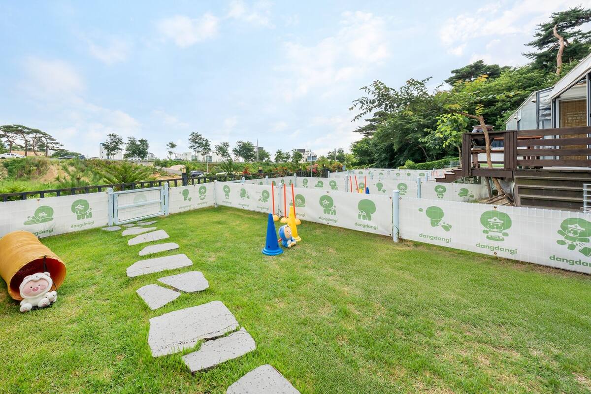 A grassy outdoor area with stepping stones, a large barrel, and a fenced playground with green signage.
