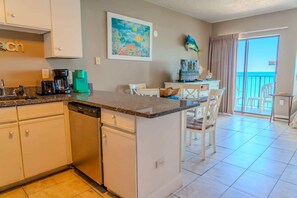 A kitchen with a dishwasher, a coffee maker, and a toaster. There is a dining area with a table and chairs. A painting is on the wall, and a fish decoration is on the ceiling.