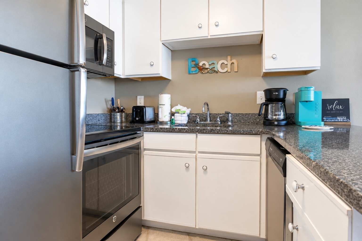 A kitchen with a stainless steel refrigerator, a microwave, a toaster, and a coffee maker. There is a sign on the wall that reads 'Beach' and a sign that says 'relax and enjoy the WiFi.'