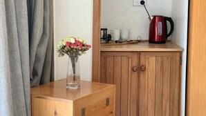 Ein hölzerner Kommode mit Blumen in einer Vase, ein roter Wasserkocher und eine blaustreifen Matratze in einem Zimmer mit einer Holztür und einem grauen Vorhang.