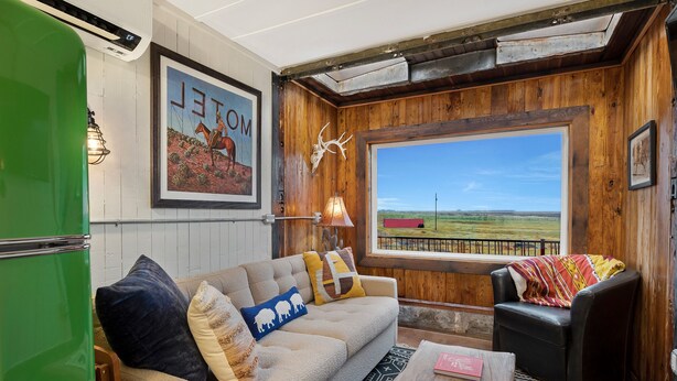A cozy living room with a sofa, a coffee table, a window with a view of the countryside, and a rustic wooden wall.