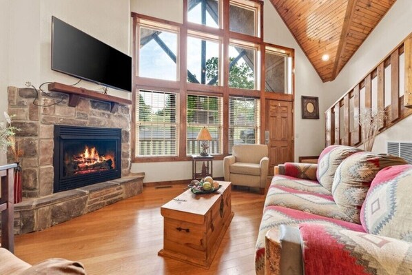 A living room with a stone fireplace, a television mounted above it, a wooden coffee table, a patterned sofa, and large windows with wooden frames. - Near Silver Dollar City! Spacious Family-sized Rental (Reeds Spring)