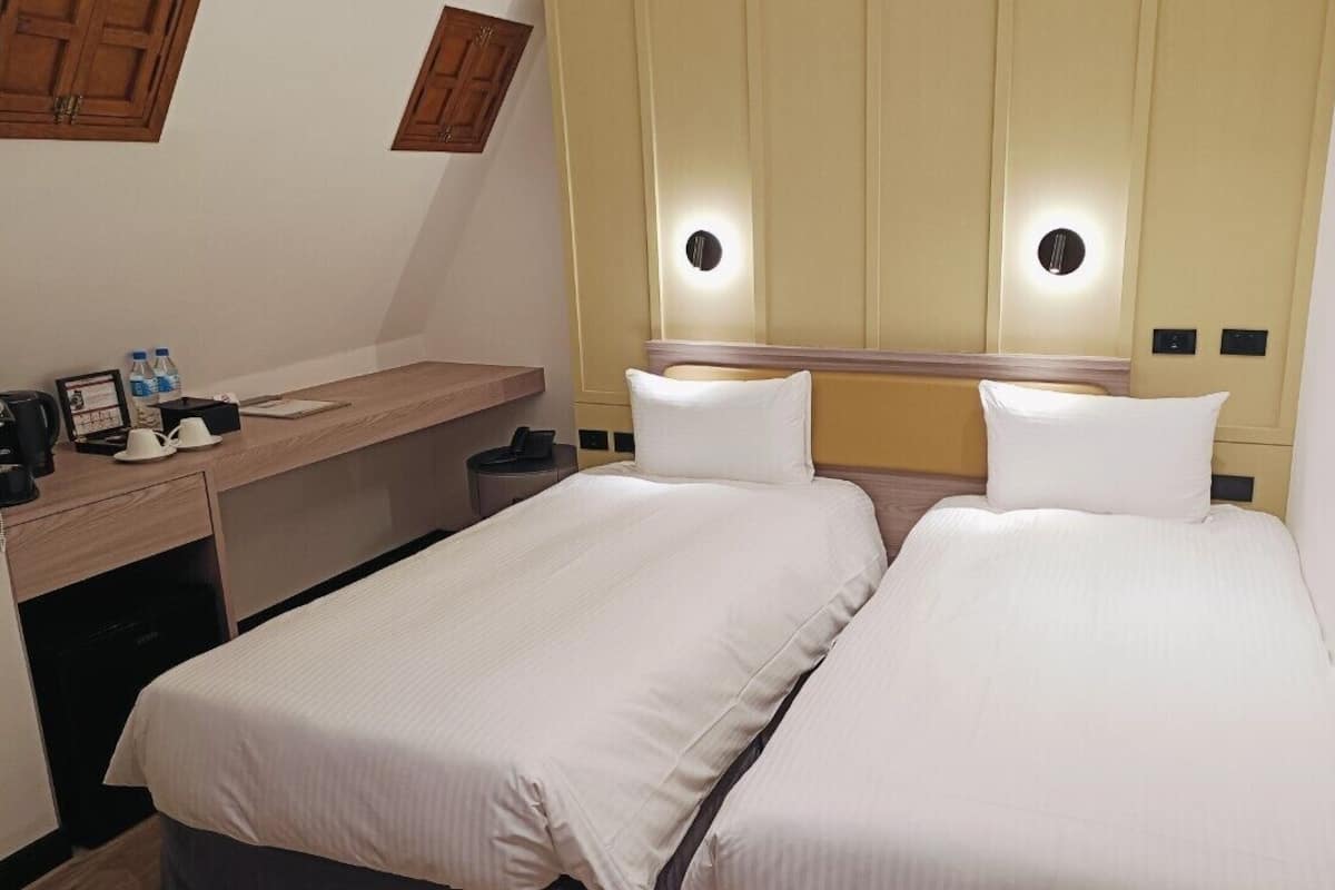 A hotel room with two single beds, a desk with a chair, a small trash bin, and a mirror above the bed.